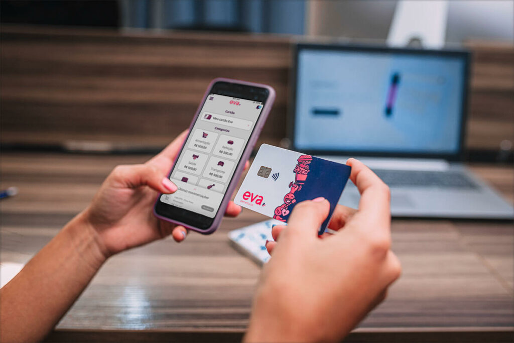 Há duas mãos na imagem, a esquerda segurando um celular exibindo o aplicativo da Eva Benefícios e a da direita segurando o cartão da Eva Benefícios