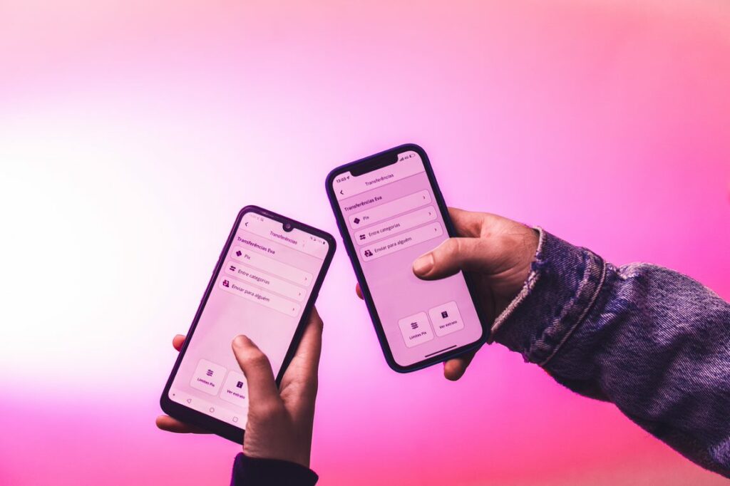 Dois celulares em frente a um fundo rosa. Ambos estão realizando transferências um para o outro.