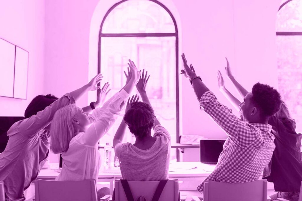 Cinco pessoas sentadas em volta de uma mesa com as mãos levantadas.