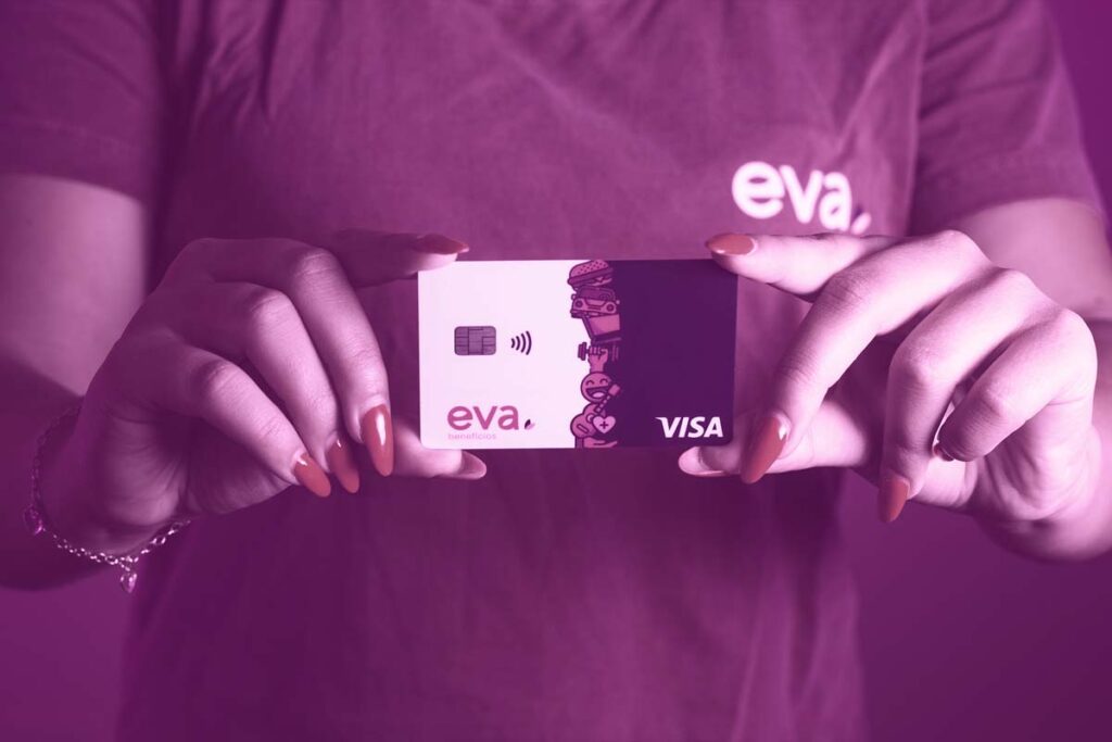 Pessoa com camiseta da 'Eva'. Nas mãos da pessoa, de unhas vermelhas, e em frente ao peito, está um cartão vale-alimentação e vale-refeição Eva.