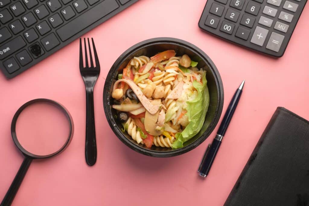 Salada de macarrão com vegetais, frango e grão-de-bico em um recipiente preto, cercada por utensílios de escritório como teclado, calculadora e caneta sobre fundo rosa.