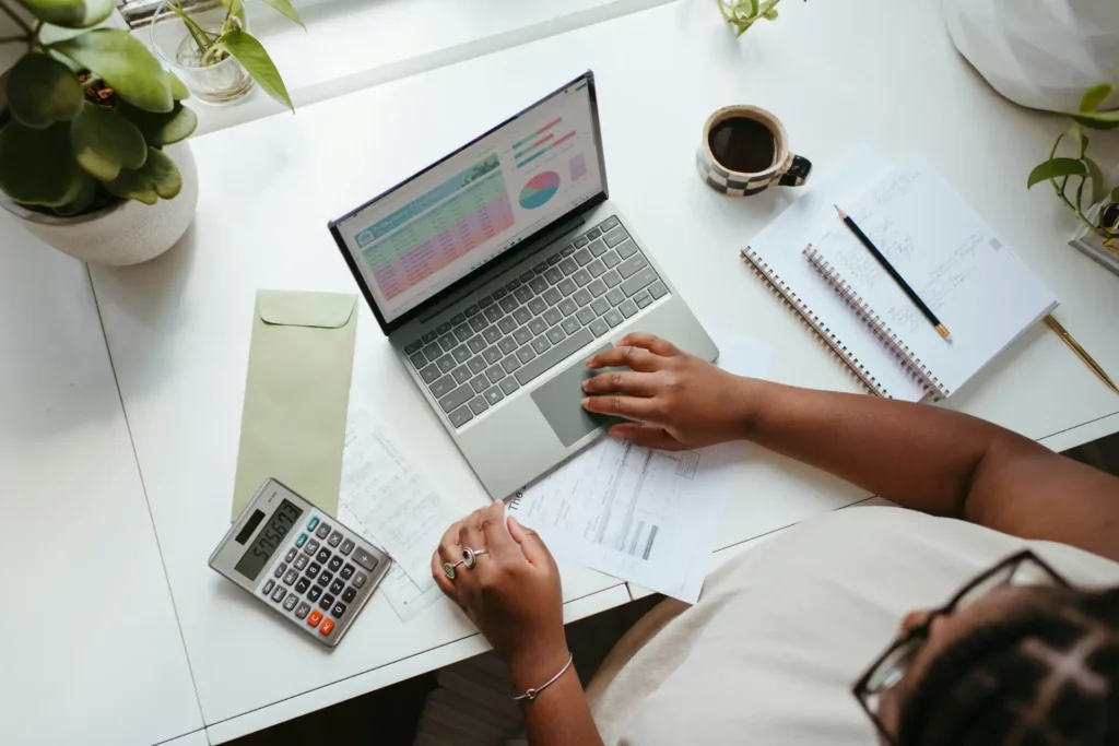 Imagem de uma mesa de trabalho mostrando uma pessoa usando um laptop, calculadora e papéis, ideal para finanças pessoais e organização. | Despesas fixas e variáveis