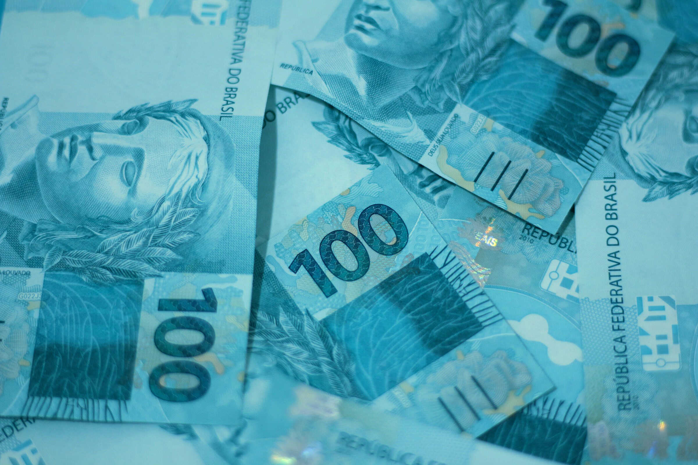Cédulas de 100 reais espalhadas sobre uma superfície, destacando o detalhe e a cor azul das notas utilizadas no Brasil.