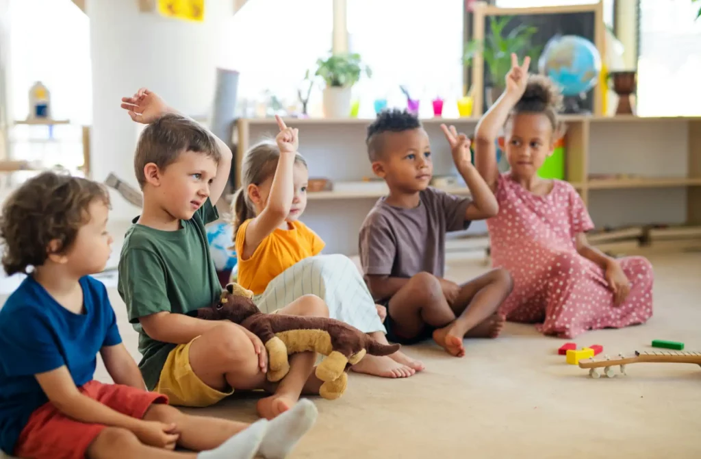 Crianças felizes levantando as mãos em sala de aula, participando de atividade lúdica com brinquedos e objetos coloridos. Ambiente acolhedor e educativo.