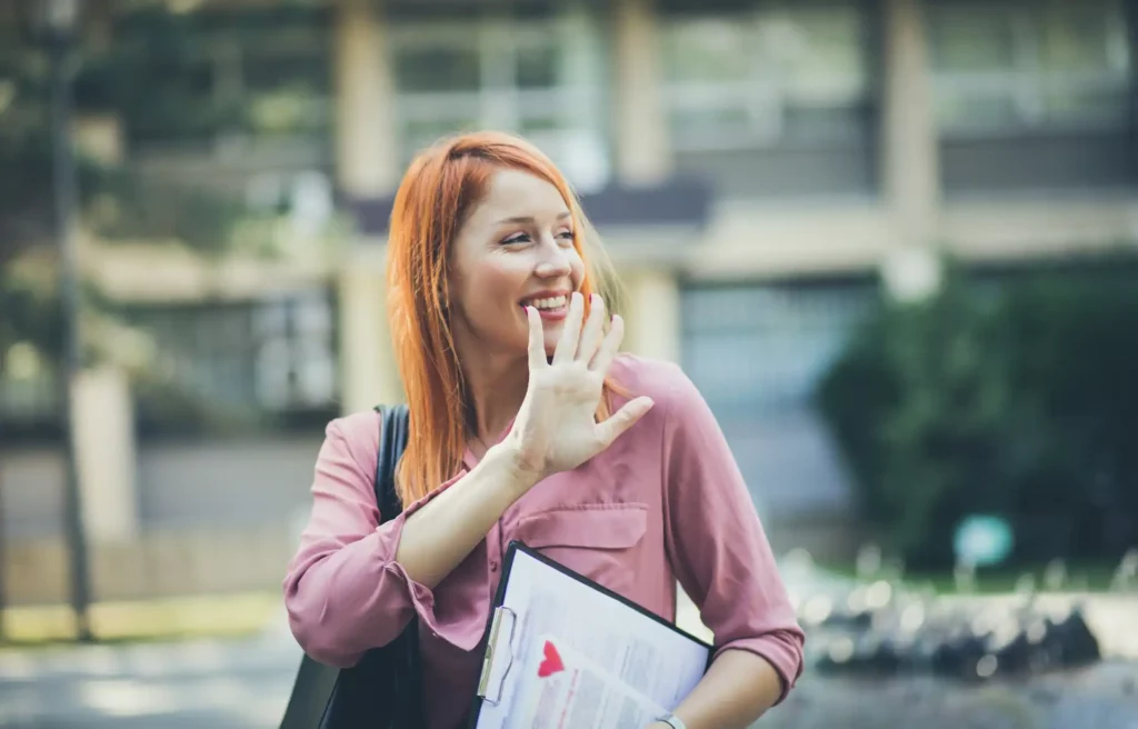 Mulher sorridente com cabelo ruivo e blusa rosa, segurando um clipboard e acenando, capturando um momento de alegria ao ar livre. | Short Friday