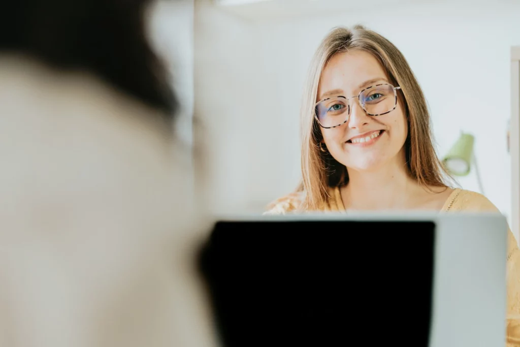 Mulher sorridente com óculos, sentada em frente a um computador, em um ambiente claro e moderno, transmitindo felicidade e confiança. | gratificação salarial