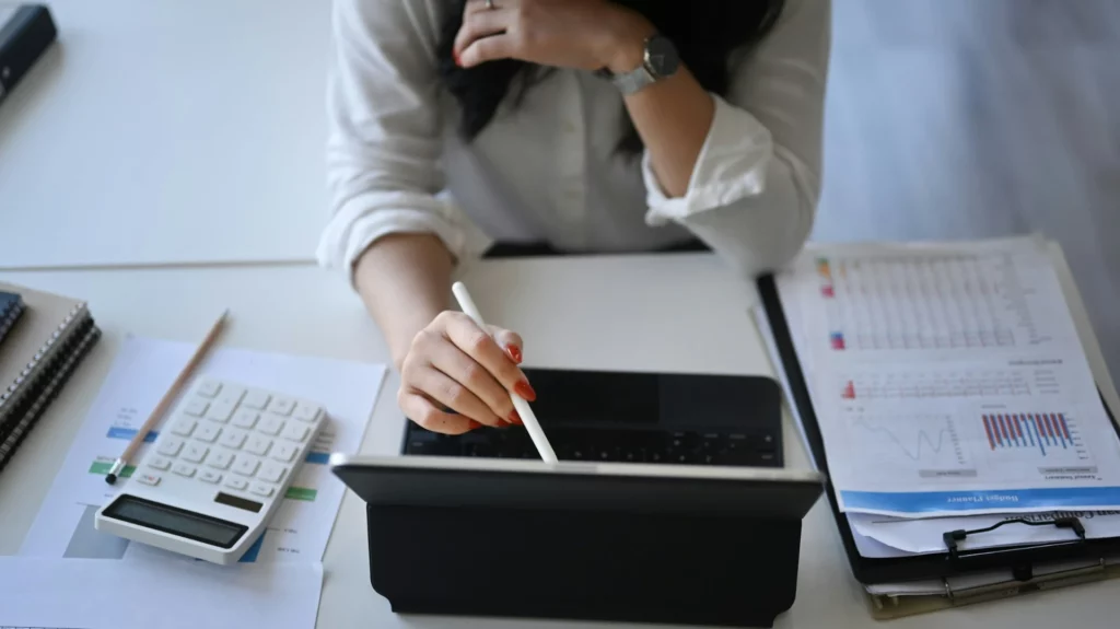 Mulher trabalhando em um tablet com calculadora e documentos, representando o ambiente de trabalho e produtividade em escritório.