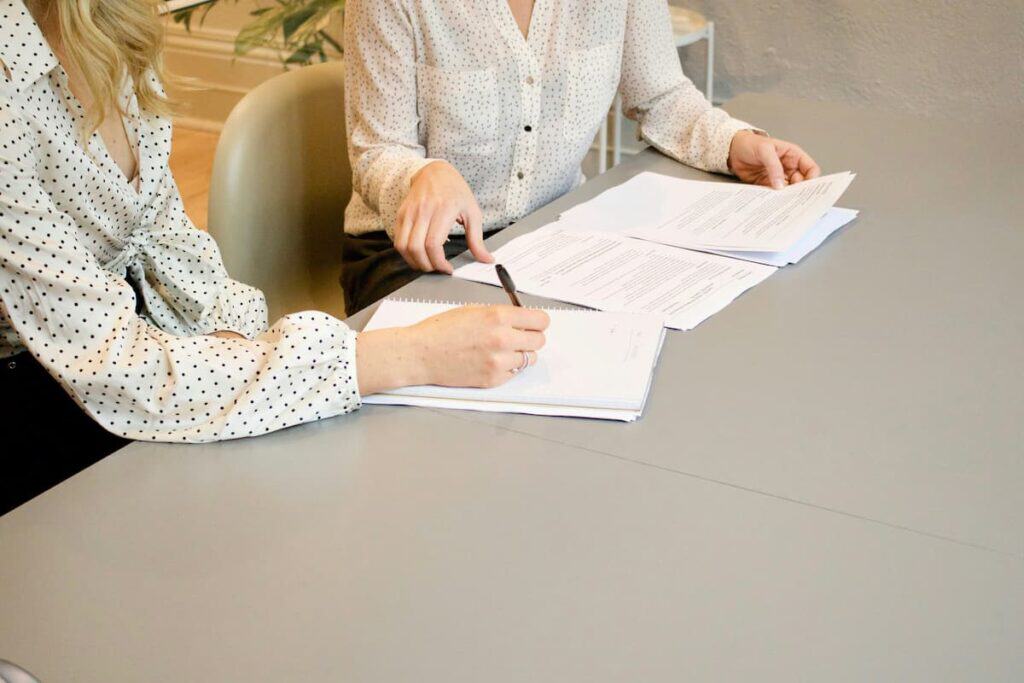Duas mulheres sentadas à mesa, assinando documentos que representam como funciona o contrato de trabalho.