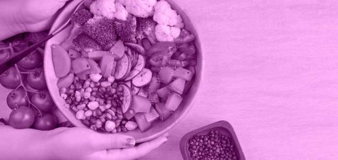 Duas mãos segurando uma cesta com frutas sobre uma mesa. Ao lado da cesta há alguns potes com outros alimentos.