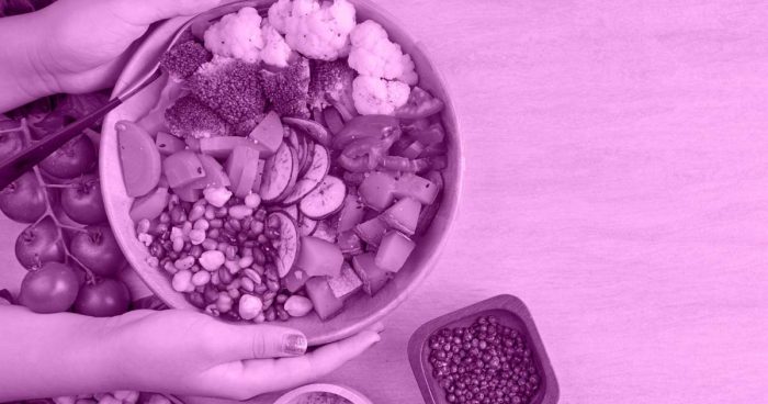 Duas mãos segurando uma cesta com frutas sobre uma mesa. Ao lado da cesta há alguns potes com outros alimentos.