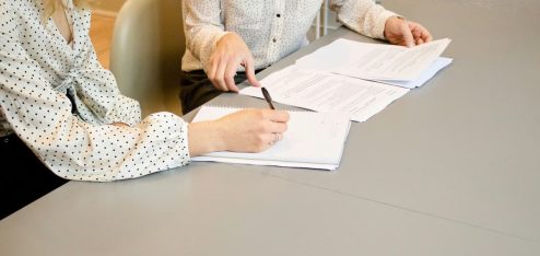 Duas mulheres sentadas à mesa, assinando documentos que representam como funciona o contrato de trabalho.