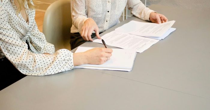 Duas mulheres sentadas à mesa, assinando documentos que representam como funciona o contrato de trabalho.