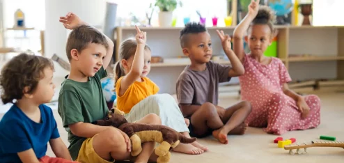 Crianças felizes levantando as mãos em sala de aula, participando de atividade lúdica com brinquedos e objetos coloridos. Ambiente acolhedor e educativo.