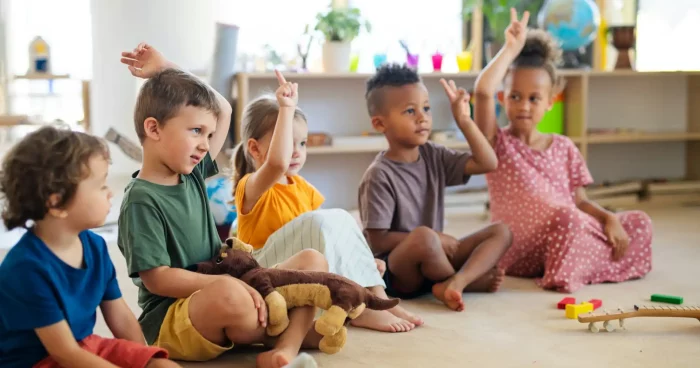 Crianças felizes levantando as mãos em sala de aula, participando de atividade lúdica com brinquedos e objetos coloridos. Ambiente acolhedor e educativo.
