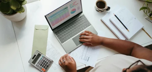 Imagem de uma mesa de trabalho mostrando uma pessoa usando um laptop, calculadora e papéis, ideal para finanças pessoais e organização. | Despesas fixas e variáveis