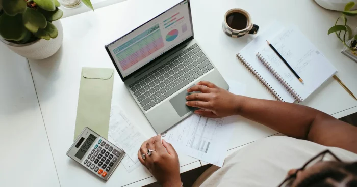 Imagem de uma mesa de trabalho mostrando uma pessoa usando um laptop, calculadora e papéis, ideal para finanças pessoais e organização. | Despesas fixas e variáveis