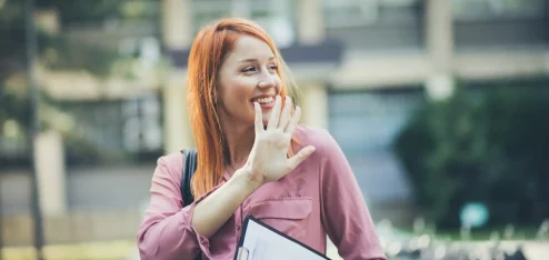 Mulher sorridente com cabelo ruivo e blusa rosa, segurando um clipboard e acenando, capturando um momento de alegria ao ar livre. | Short Friday