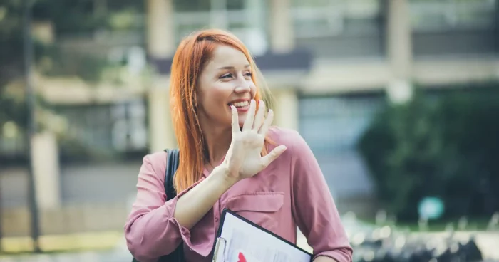 Mulher sorridente com cabelo ruivo e blusa rosa, segurando um clipboard e acenando, capturando um momento de alegria ao ar livre. | Short Friday