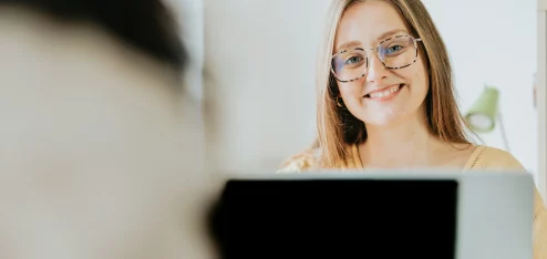 Mulher sorridente com óculos, sentada em frente a um computador, em um ambiente claro e moderno, transmitindo felicidade e confiança. | gratificação salarial