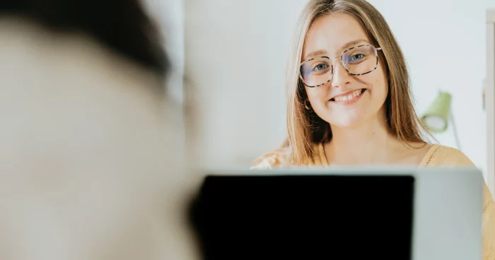 Mulher sorridente com óculos, sentada em frente a um computador, em um ambiente claro e moderno, transmitindo felicidade e confiança. | gratificação salarial