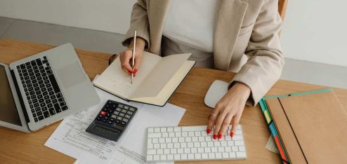 Pessoa analisando documentos financeiros com laptop e calculadora em mesa de escritório, representando a prestação de contas empresarial.