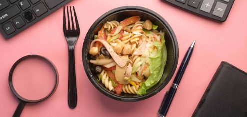 Salada de macarrão com vegetais, frango e grão-de-bico em um recipiente preto, cercada por utensílios de escritório como teclado, calculadora e caneta sobre fundo rosa.