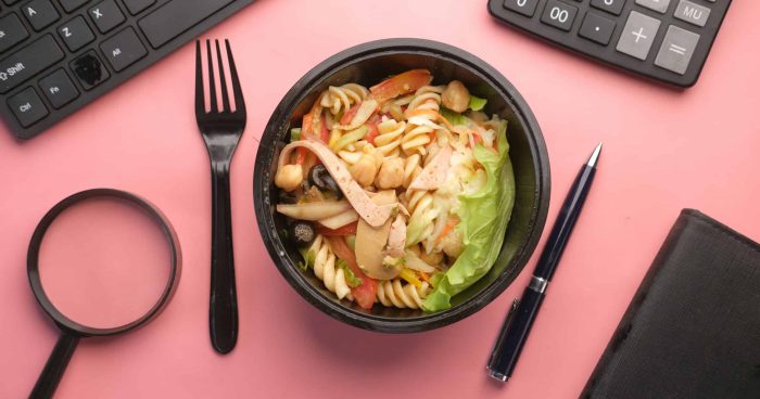 Salada de macarrão com vegetais, frango e grão-de-bico em um recipiente preto, cercada por utensílios de escritório como teclado, calculadora e caneta sobre fundo rosa.